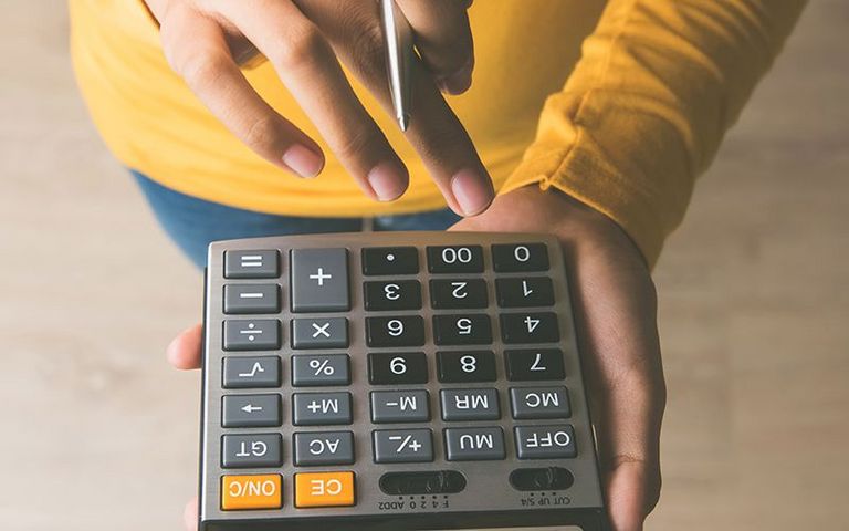 Eine Frau in einem gelben Pullover hält einen Taschenrechner in der Hand und tippt Zahlen ein.